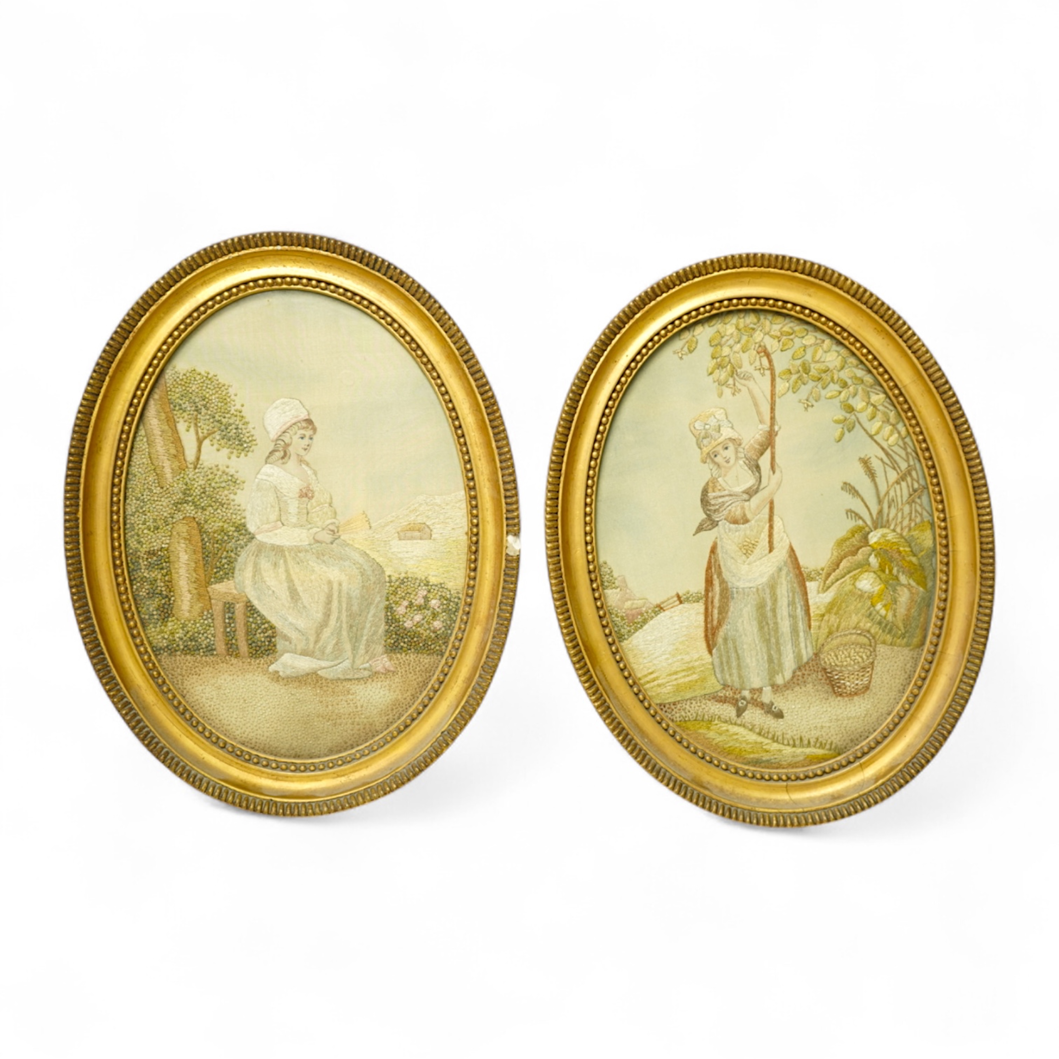A pair of early 20th century small gilt oval embroideries, of a girl gathering fruit from a tree and a seated lady in a landscape, worked in stem stitch and decorative knotting. 29cm long including gilt frames. Condition
